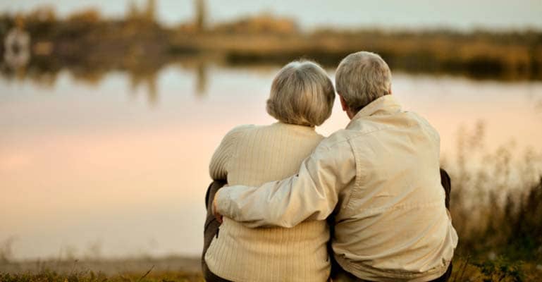 Elderly couple embracing while seated next to a lake at sunrise