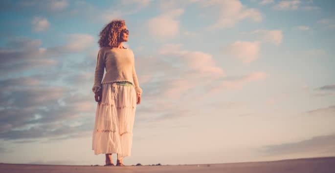 Curly-haired woman with sunglasses looks at sunset while on the beach with sky behind her