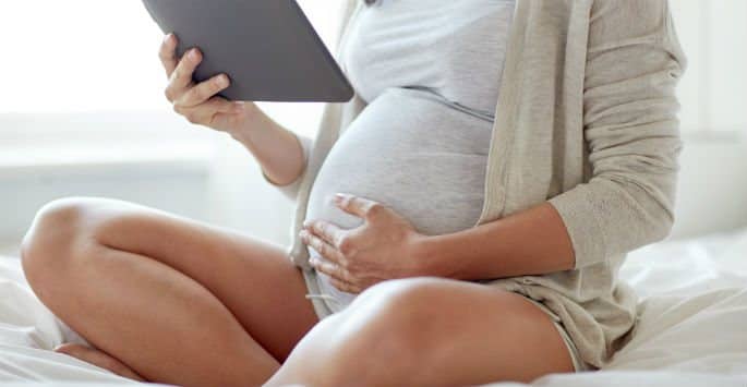 A pregnant woman with her legs crossed sitting on her bed while viewing her tablet device