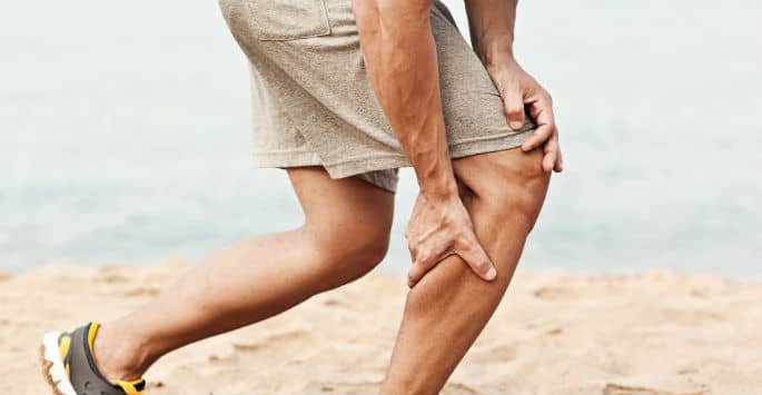 A man in shorts grabbing is legs as he runs on the beach