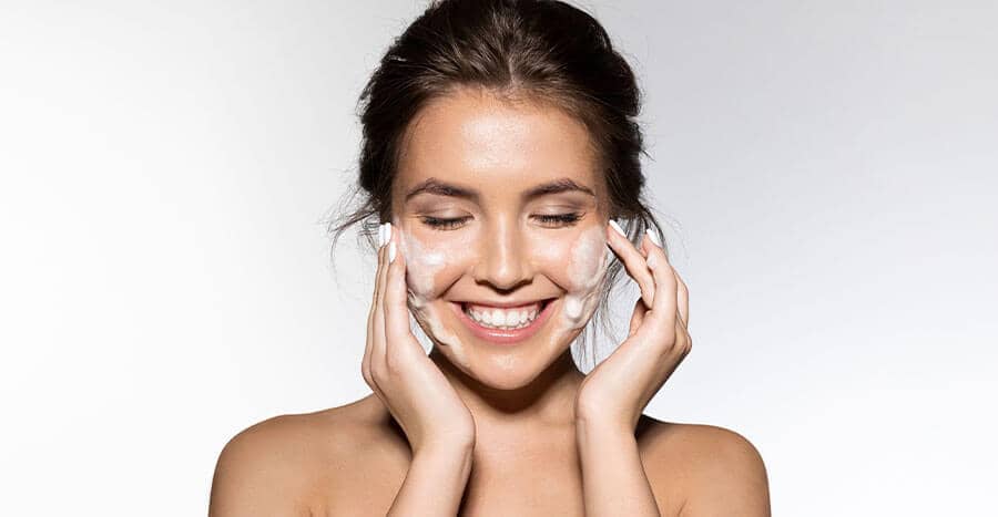 Smiling female with her hair pulled back as she applies facial cleanser to her face
