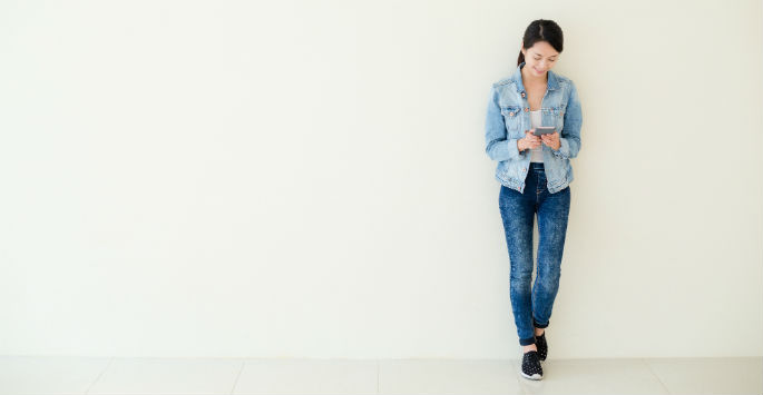 An Asian woman wearing jeans leaning against a wall while viewing smartphone
